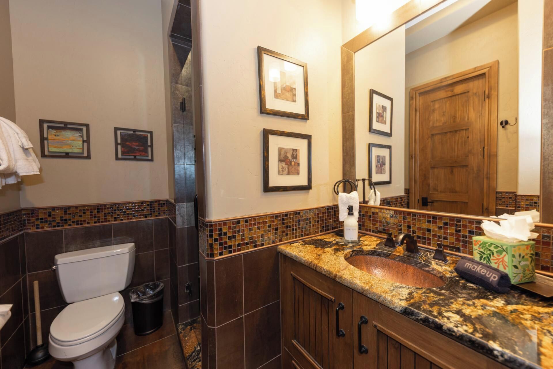 A bathroom with a toilet, sink, and mirror. There are framed pictures on the wall and a towel rack.