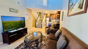 A living room with a TV, sofa, and coffee table. There's a staircase leading to a kitchen and a dining area.