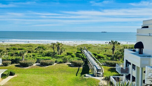 A coastal view with a beach, a white building with a balcony, and a pathway leading to the beach.