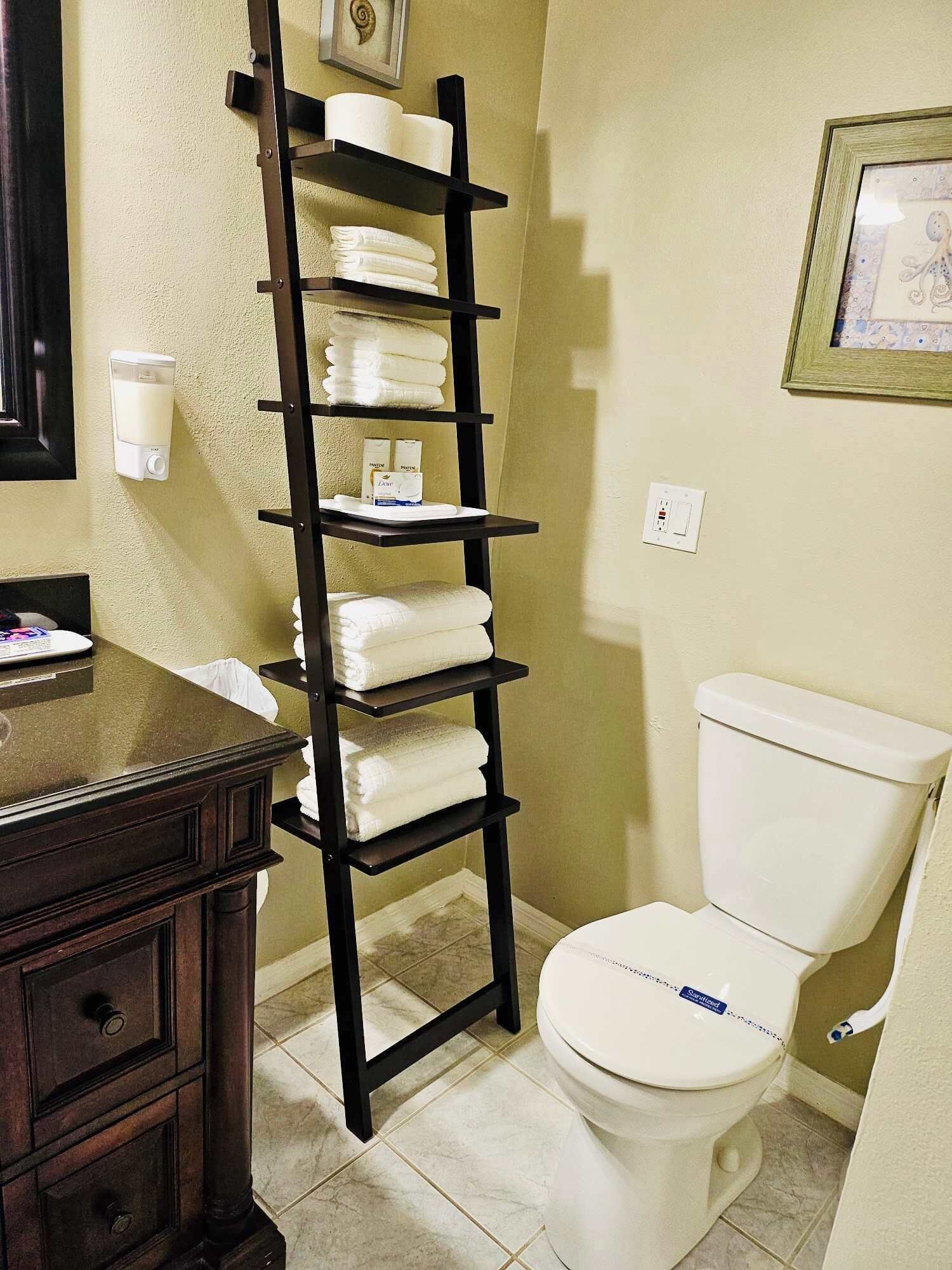 A bathroom with a toilet, a shelf with folded towels, a soap dispenser, and a framed picture on the wall.