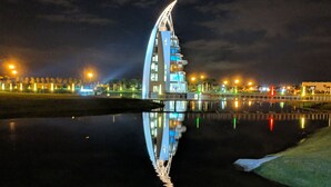 Un edificio moderno con un diseño curvo distintivo, reflejado en una superficie acuática tranquila, rodeado por una zona urbana bien iluminada.