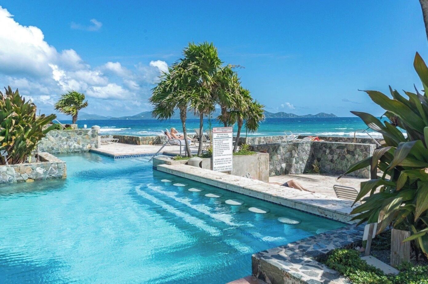 Una piscina con vistas al océano, rodeada de palmeras y un muro de piedra.
