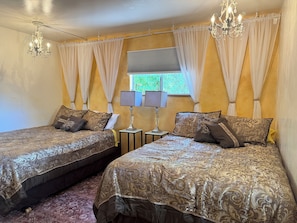 A room with two beds, a window with curtains, a chandelier, and a lamp.