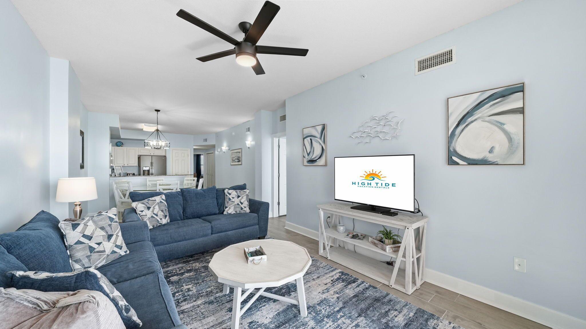 A modern living room with a sofa, coffee table, and TV. There is a ceiling fan, artwork on the walls, and a kitchen in the background.