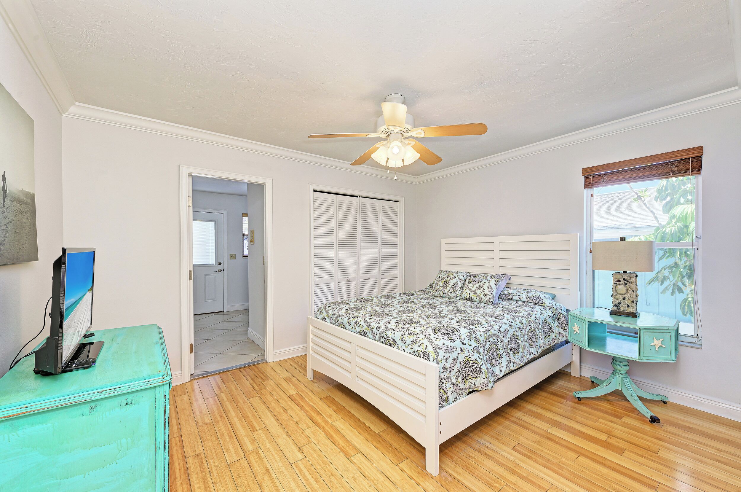 Une chambre à coucher avec un lit, un ventilateur de plafond, un bureau avec une lampe et une fenêtre avec des stores.