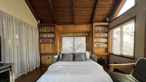 Ein gemütliches Schlafzimmer mit einem großen Bett, Holzregalen, einem Fenster mit Jalousien und einem grünen Sessel.