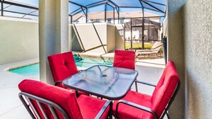 Ett takförsett utomhusområde med sittplatser, en pool, röda stolar och ett bord i glas.
