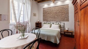Une chambre avec un mur en pierre, un plafond en bois, un lit recouvert d’une couverture verte, une table de chevet, une armoire en bois et une table à manger ornée d’un vase de fleurs.