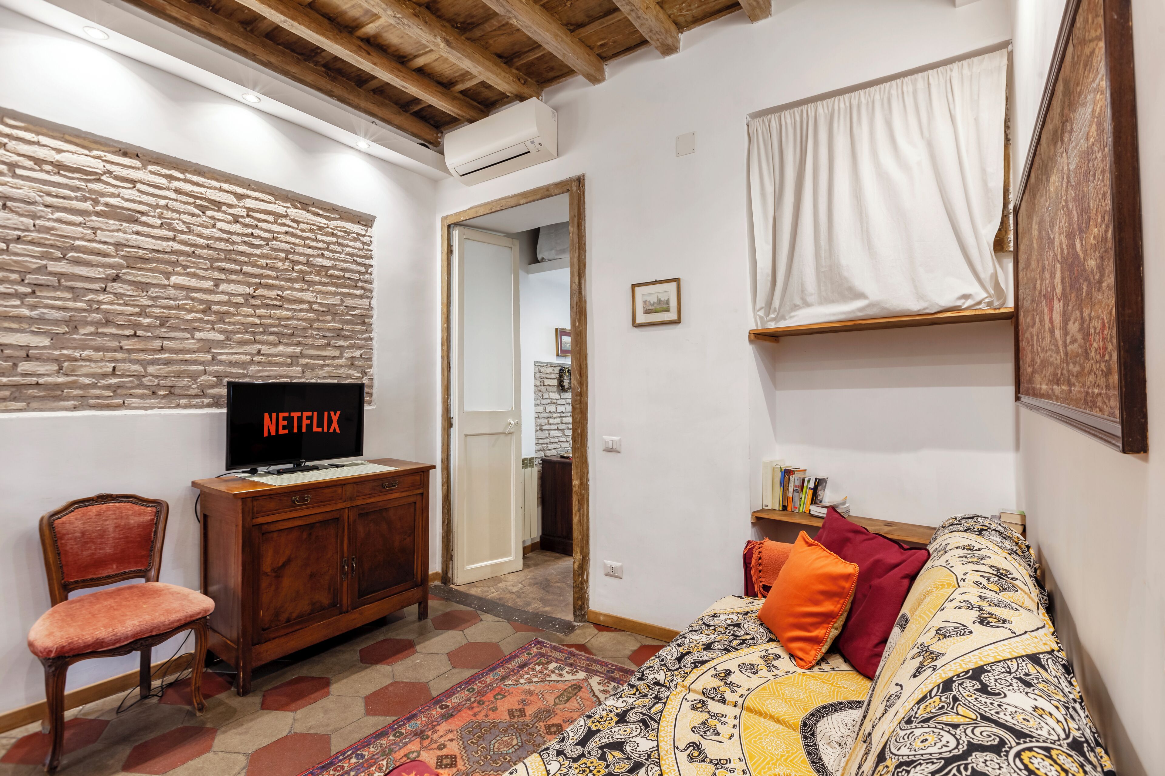A living room with a television displaying 'NETFLIX,' a wooden cabinet, a patterned sofa, and a brick wall.