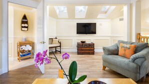 Un salón con piso de madera, un sofá gris con un cojín naranja, un televisor montado en la pared, un aparador de madera y una orquídea en maceta.