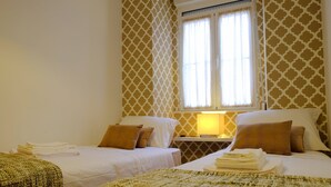A hotel room with two beds, a window with curtains, a bedside table with a lamp, and a woven rug. - Travessa House I - Graça. Lisbon Historic Center (Lisbon)