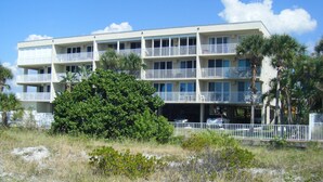 A multi-story hotel with balconies, a parking garage, and a large tree in the foreground. - Special Nov 14-21, 2026 - IRB Direct Beach Front Corner Condo (Indian Rocks Beach)