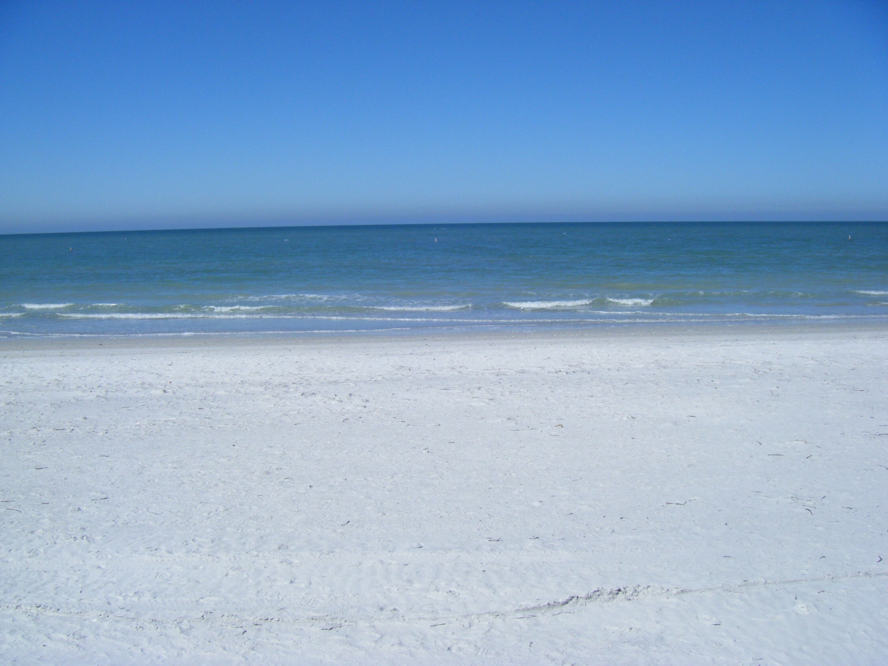 A wide, sandy beach with gentle waves lapping at the shore under a clear blue sky.