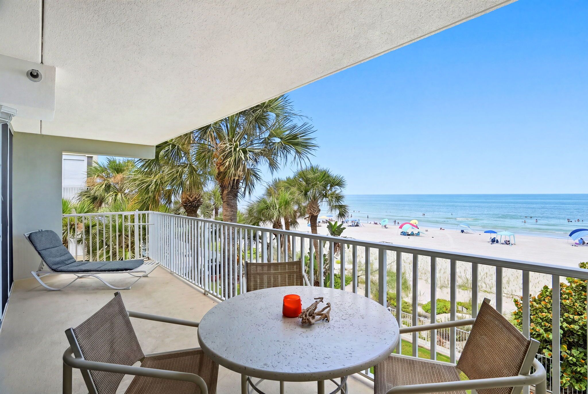 A balcony with a table and chairs, overlooking a beach with palm trees and the ocean.