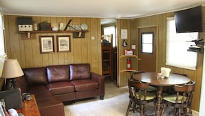 A living room with a brown leather sofa, wooden walls, a dining table with chairs, and a television mounted on the wall. - Walk to White Oak Canyon Trail in Shenandoah National Park (Syria)