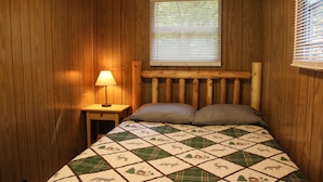 A bedroom with wooden walls, a bed with a quilt, a nightstand with a lamp, and a window with blinds. - Walk to White Oak Canyon Trail in Shenandoah National Park (Syria)