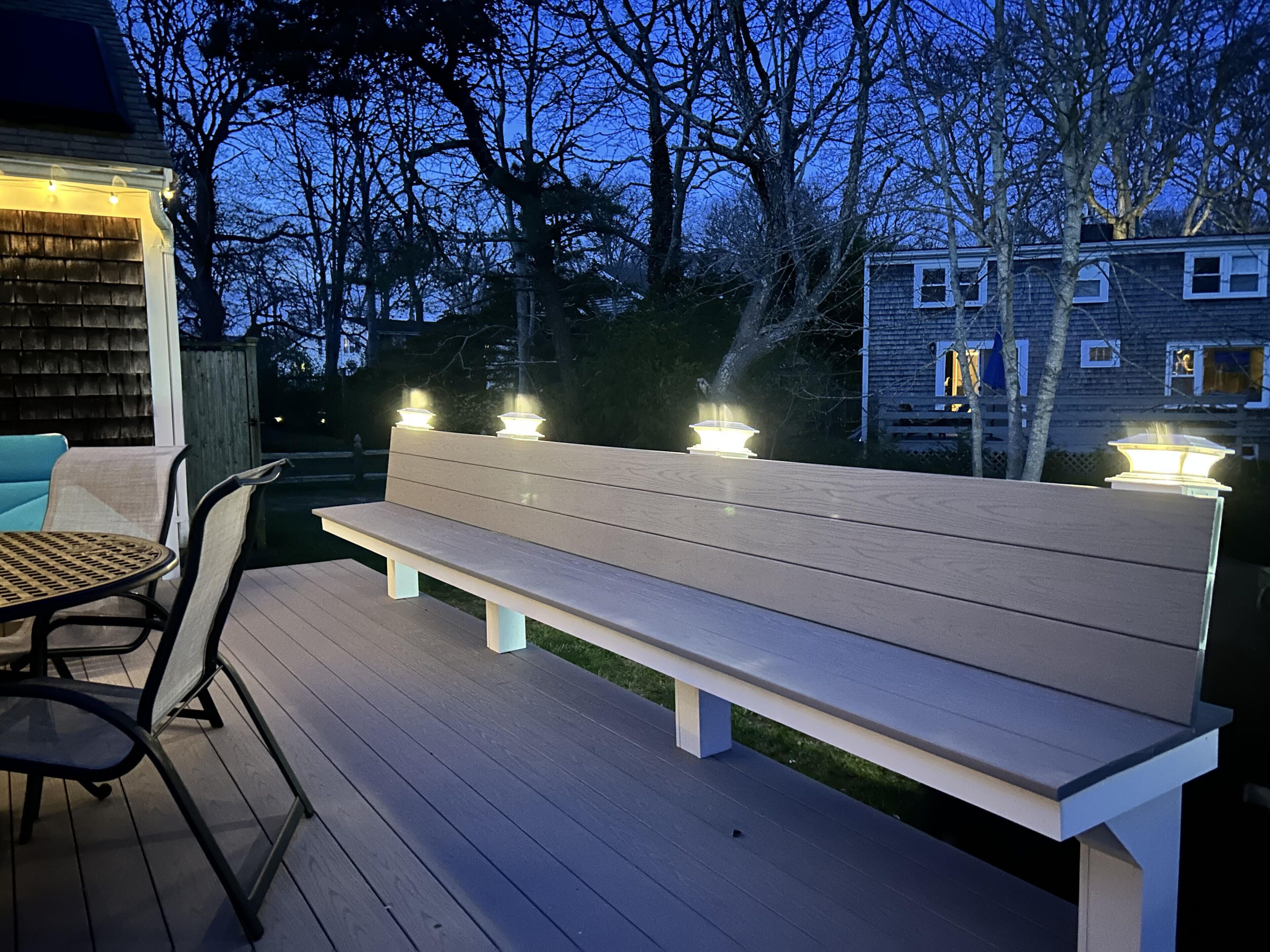 An outdoor seating area with a bench and chairs, surrounded by trees and a house in the background.