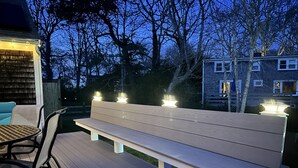 Un espace extérieur aménagé avec un banc et des chaises, entouré d’arbres et avec une maison en arrière-plan.