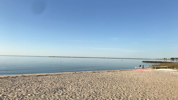 Une plage avec une eau calme, un ciel dégagé et une jetée qui se profile à l’horizon.