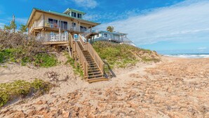 A beachfront property with a staircase leading to a balcony, a sandy beach, and the ocean in the background.
