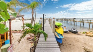 A pier leading to a beach with a row of kayaks and jet skis, palm trees, and a wooden structure with a sign.