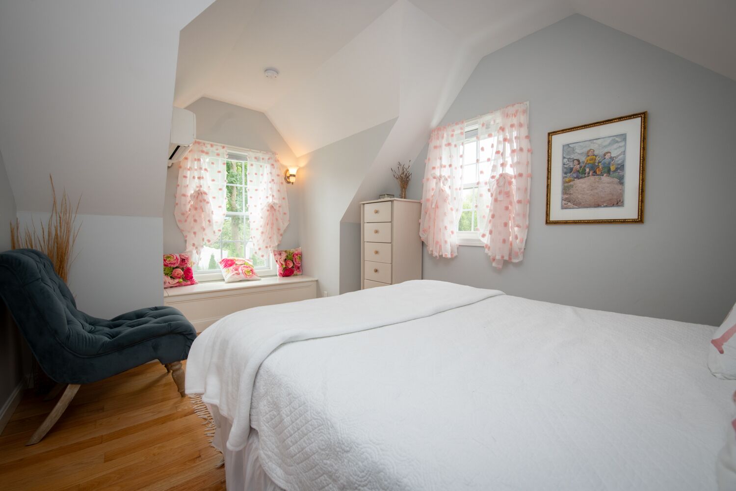 Quarto com cama grande, uma cadeira, cômoda e duas janelas com cortinas.
