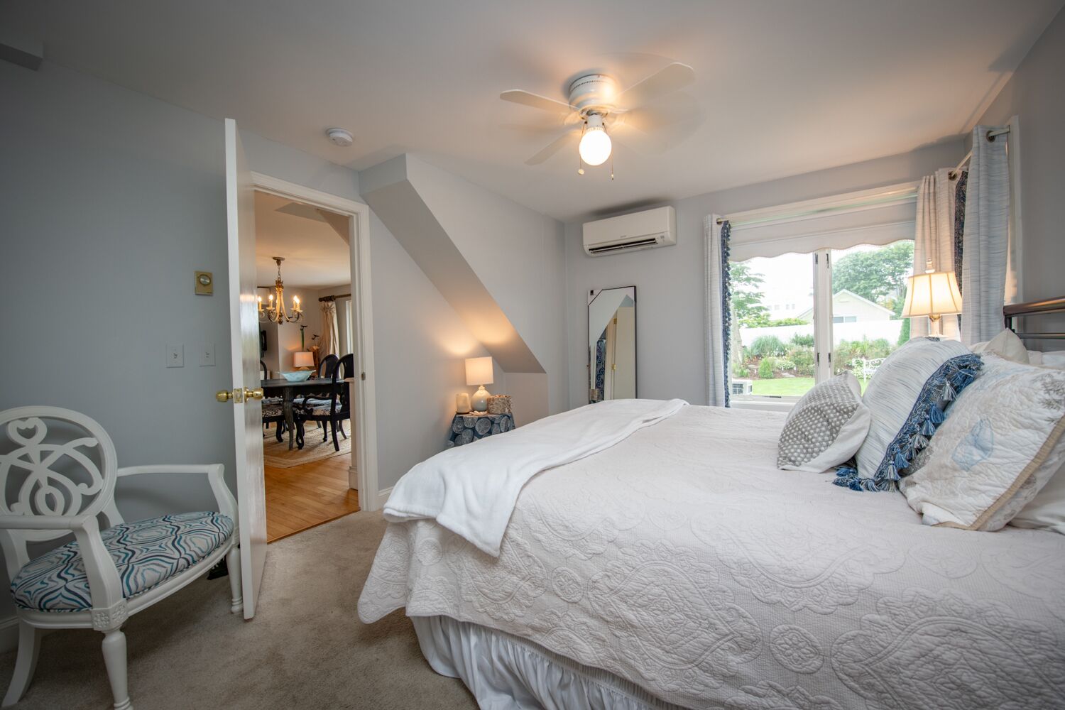 A bedroom with a large bed, a ceiling fan, an air conditioning unit, a dining area with a chandelier, and a view of the outdoors.