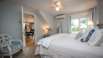 A bedroom with a large bed, a ceiling fan, an air conditioning unit, a dining area with a chandelier, and a view of the outdoors.