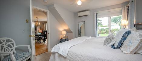 A bedroom with a large bed, a ceiling fan, an air conditioning unit, a dining area with a chandelier, and a view of the outdoors.