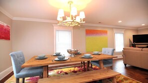A dining area with a wooden table, chairs, and a chandelier. There is a sofa and a TV in the adjacent living area. - Luxury New Home: Ocean and City Light View Home + Steps to Restaurants sleeps 6 (Carolina Beach)