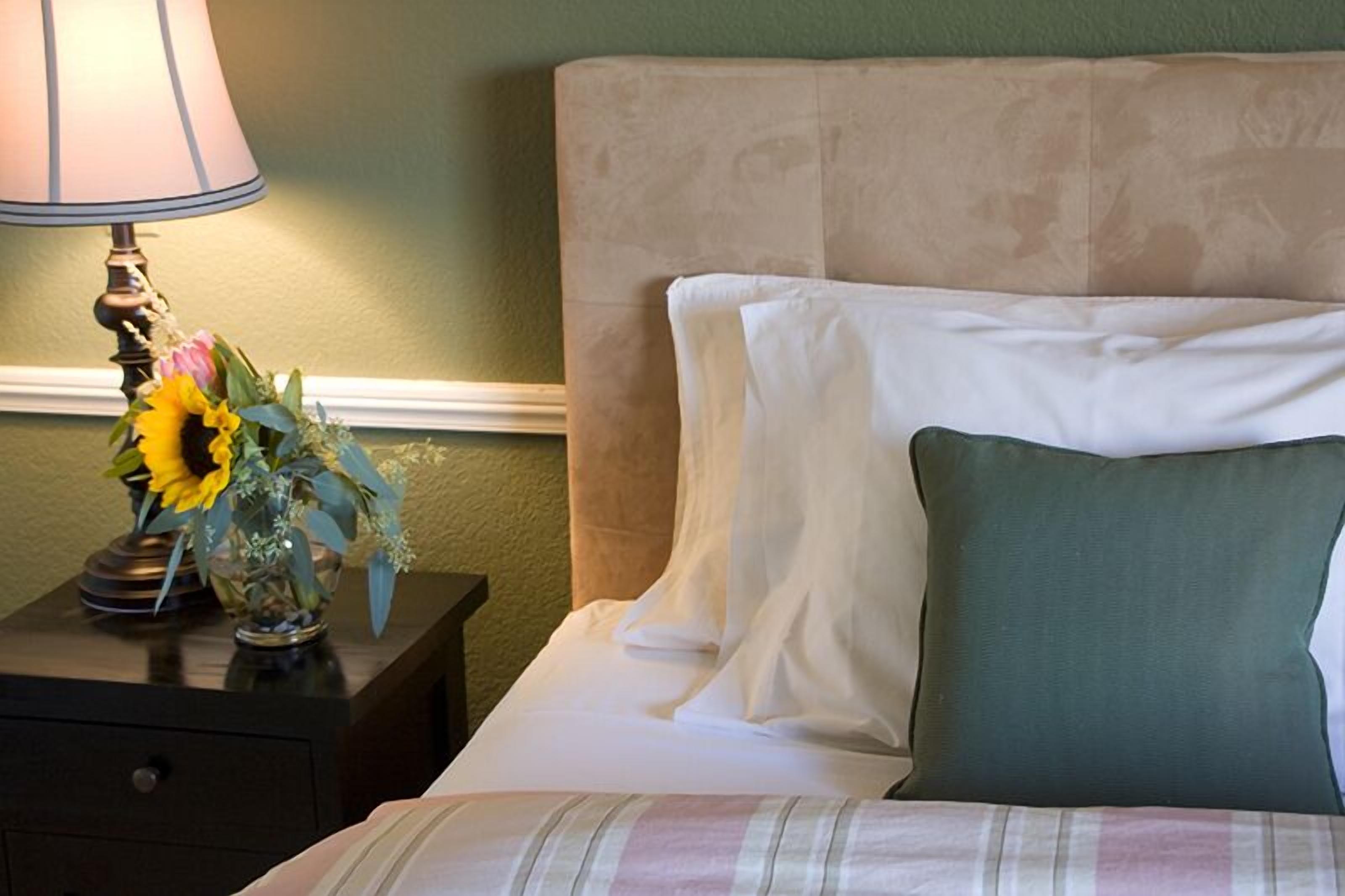 Uma cama bem arrumada com almofadas brancas e verdes, um cobertor listrado, uma mesa de cabeceira com abajur e um vaso de flores.