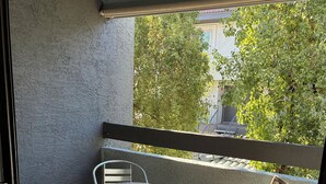 Un balcón con una mesa de metal y dos sillas de madera, una silla de metal, y vistas a árboles y un edificio al fondo.