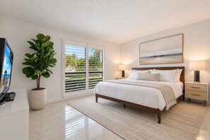 A bedroom with a bed, bedside tables, a TV, a large window with blinds, and a potted plant.