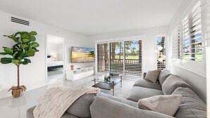 A modern living room with a grey sofa, a glass coffee table, and a large window with blinds.