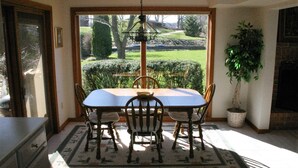 Zona pranzo con tavolo e sedie, un lampadario, una pianta in vaso e vista su un giardino attraverso una grande finestra.