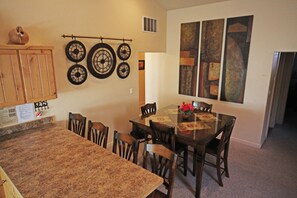 A dining area with a large wooden table, six chairs, and a wall-mounted clock. There are abstract paintings on the wall and a wooden cabinet with a small decorative item on top. - Southwest Charm and Amazing Views, just minutes from town! (Moab)