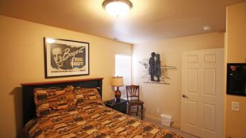 Ein Schlafzimmer mit einem gemusterten Bettüberwurf, einem gerahmten Bild an der Wand, einem Sessel, einem Nachttisch, einer Lampe und einer Tür.