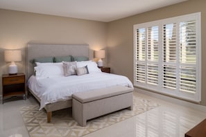 A bedroom with a bed, nightstands, a bench, and a large window with blinds.