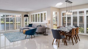 A spacious living room with a sofa, chairs, a dining table set for six, and large windows with blinds.