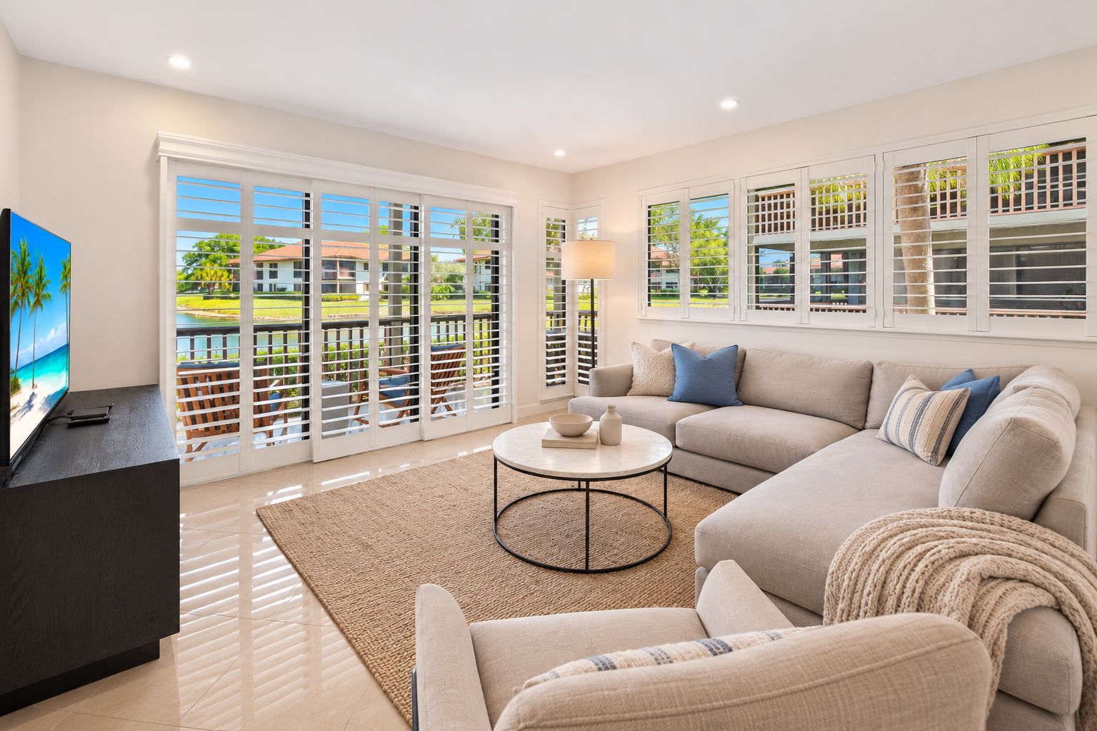 A modern living room with a sofa, coffee table, and TV, large windows with blinds, and a view of a pool and greenery.
