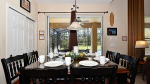 Une salle à manger avec une table dressée pour quatre personnes, des chaises, un lustre et une vue sur l’extérieur, visible à travers la porte vitrée.