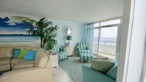 Ein Zimmer mit einem Wandgemälde zum Thema Strand, einer großen Kübelpflanze, gestreifter Ohrensessel, einem Sofa und Meerblick durch große Fenster.