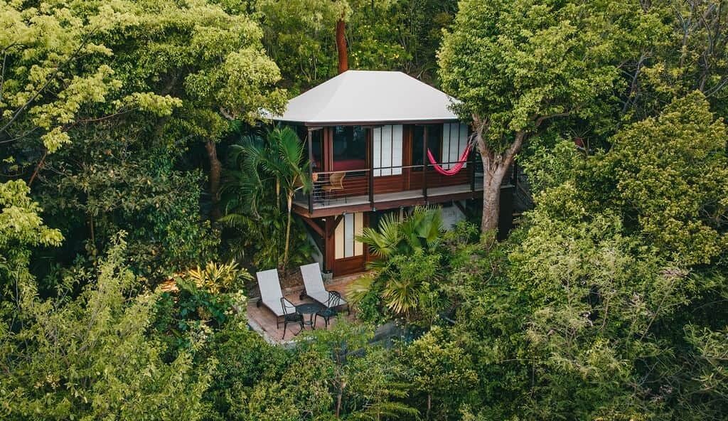 A two-story house surrounded by dense greenery, with a balcony and outdoor seating area.