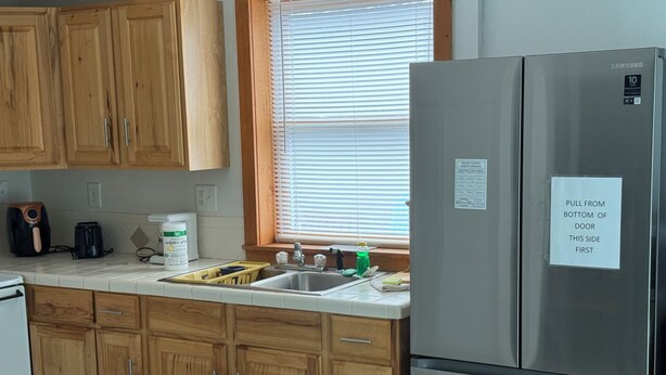 A kitchen with wooden cabinets, a stainless steel refrigerator, a window with blinds, and a sink with various items on the counter. - Apartment in town (Franklin)