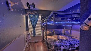 Una cama nido con edredón con tema de galaxia, una ventana con persianas y un techo decorado con estrellas.