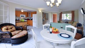 Un coin repas avec une table ronde dressée pour quatre personnes, un canapé en osier et une cuisine en arrière-plan.