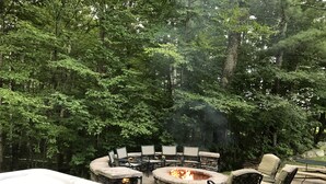 An outdoor patio with a stone fire pit, seating, and a hot tub surrounded by lush greenery.