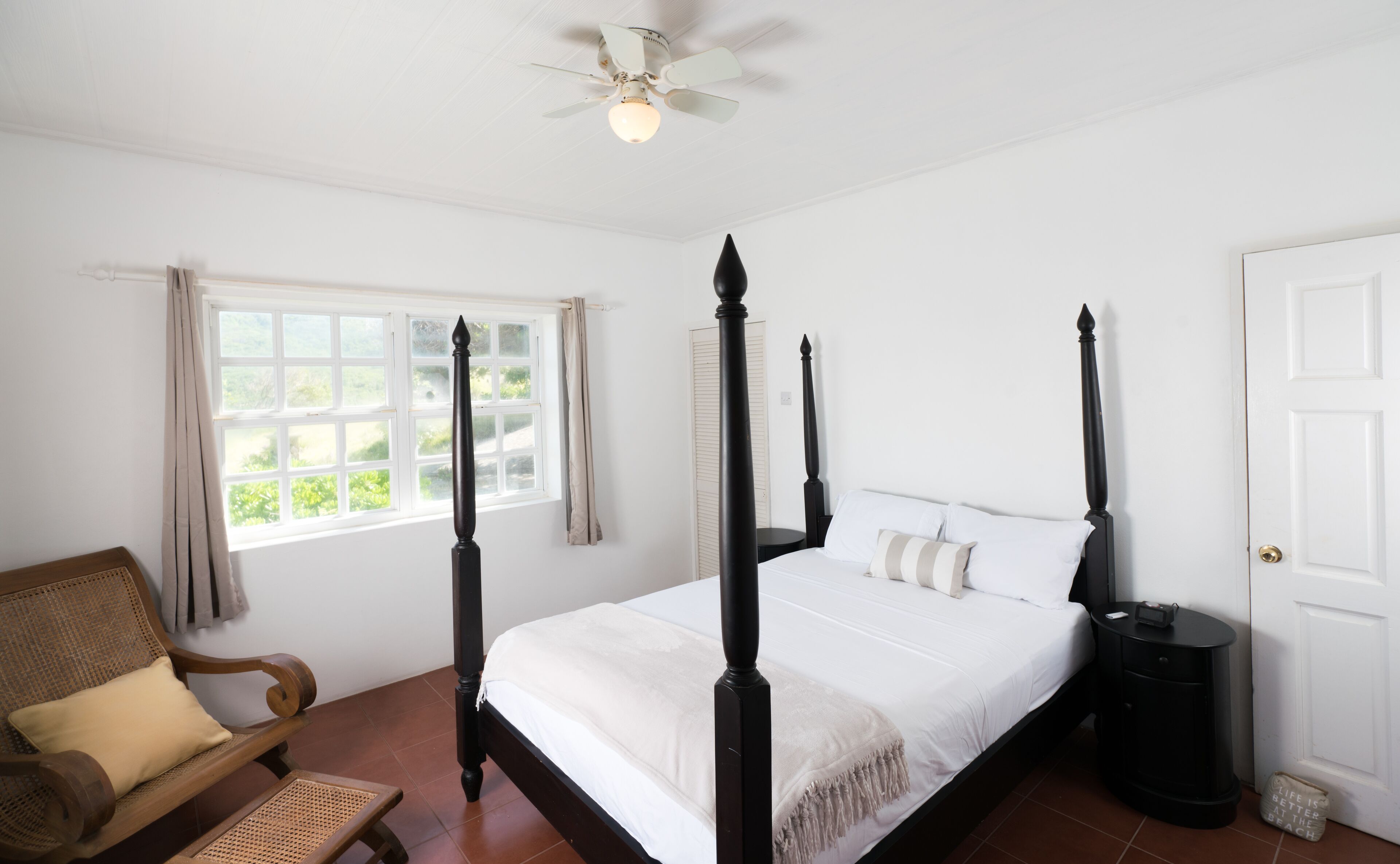 A four-poster bed with a white bedspread and multiple pillows, a wooden chair with a cushion, a ceiling fan, and a window with curtains.