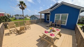 A deck with wooden furniture, a table with fruit and drinks, and a blue building in the background.
