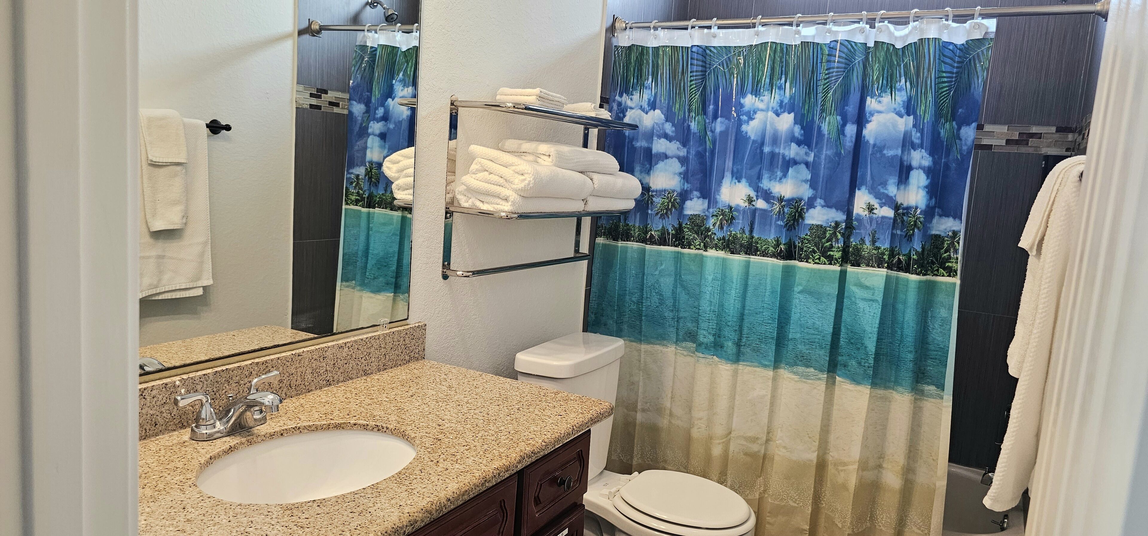 Une salle de bain avec un lavabo sur un plan de travail en granit, des toilettes, un porte-serviettes avec des serviettes pliées et un rideau de douche représentant une plage tropicale.